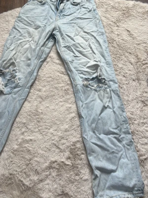 Ljusblå ripped jeans från Gina Tricot - Säljer ett par ljusblå jeans från Gina Tricot med slitna hål på båda knäna. Klassisk femficksmodell med dragkedja och knapp. Jeansen har en avslappnad passform och är gjorda i bomullsdenim. Perfekta för en trendig och avslappnad look.