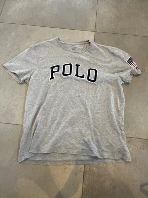 Grå POLO t-shirt med tryck - Snygg grå t-shirt från Polo Ralph Lauren med stort svart POLO-tryck på bröstet och amerikansk flagga på ärmen. Klassisk passform och rund halsringning. Perfekt för en avslappnad och sportig stil.