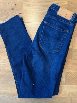 Mörkblå jeans från Tiger of Sweden - Snygga mörkblå jeans från Tiger of Sweden med klassisk femficksmodell och råa, avklippta benslut. Jeansen har en rak passform och är tillverkade i mjuk denim med läderpatch bak i midjan.