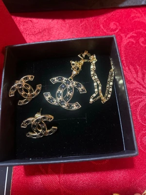 Chanel halsband med CC-logga i guld - Säljer ett elegant set halsband från Chanel med den ikoniska CC-loggan i guldtonad metall och svarta detaljer. Kedjan är tunn och stilren, och hänget är dekorerat med små stenar för extra bling. Perfekt för dig som vill ha en lyxig accessoar med statement-känsla. Matchande örhängen för hål i öronen ingår