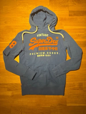 Blå Superdry hoodie med tryck - Snygg blå hoodie från Superdry med stor orange och vit logga samt japansk text på bröstet. Hoodien har vita dragsnören, känguruficka och ribbade muddar. Storlek S, inga defekter som ni ser på bilden. Hör av dig vid frågor eller prisförslag!