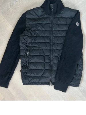  Moncler Cardigan  - Snygg svart dunväst från Moncler med quiltad front och stickade ärmar. Västen har dragkedja framtill, två fickor med dragkedja och en liten Moncler-logga på ena ärmen. Perfekt för lager-på-lager och streetstyle.