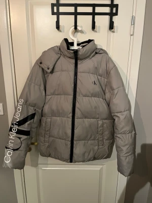 Grå pufferjacka från Calvin Klein - Snygg grå pufferjacka från Calvin Klein Jeans med svart dragkedja, huva och logga på bröstet. Jackan har stora fickor och coolt tryck på ena ärmen. Perfekt för kalla dagar och riktigt trendig look.