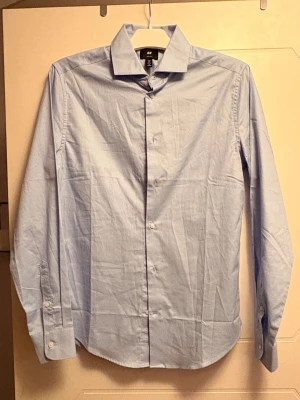 Ljusblå skjorta från H&M - Klassisk ljusblå skjorta från H&M med lång ärm och knappar framtill. Skjortan har en stilren design med krage och är tillverkad i bomull, perfekt för en clean och enkel look.