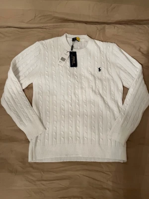 Vit kabelstickad tröja Polo Ralph Lauren - Kabelstickad vit tröja från Polo Ralph Lauren med klassisk rund halsringning och svart broderad logga på bröstet. Tröjan har ribbade muddar vid ärmslut och nederkant, och är gjord i mjukt material som ger en clean och stilren look.
