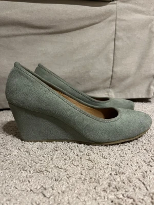 Grågröna kilklackade pumps - Snygga pumps i grönt mockaliknande material med kilklack. Skorna har rundad tå och en stilren, enkel design utan dekorationer. Använda en gång på min bal.