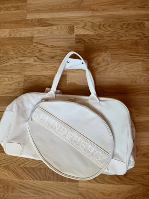 Vit sportbag från J.Lindeberg - Snygg vit sportbag från J.Lindeberg med stort huvudfack, avtagbar axelrem och praktiska handtag. Väskan har en rund ficka med dragkedja och tydlig logga på sidan. Tillverkad i slitstarkt material, perfekt för gym eller resa. Nypris 2400-2800