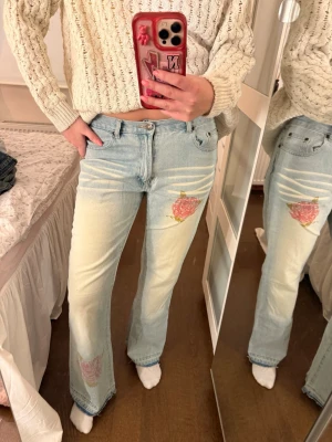 Ljusblå jeans med rosapatch - Unika ljusblå jeans med broderade rosa rosor på låret och vid benslutet. Jeansen har klassisk femficksmodell och är tillverkade i denim. Perfekta för dig som vill sticka ut med coola detaljer och en avslappnad vibe.