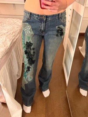 Blå jeans med gröna pärlapplikationer - Unika blå jeans med coola gröna och svarta pärlapplikationer samt broderier på ena benet och bakfickan. Jeansen har klassisk femficksmodell och stängs med knapp och dragkedja. Perfekta för dig som vill sticka ut med din stil.