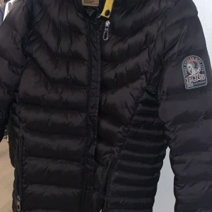 Svart dunjacka från Parajumpers - Snygg svart dunjacka från Parajumpers med gul detalj vid kragen och patch på ärmen. Jackan har quiltad design, dragkedja framtill och hög krage. Perfekt för kalla dagar och riktigt varm tack vare dunfyllningen.