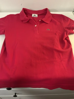 Rosa pikétröja från Lacoste - Säljer en klassisk rosa pikétröja från Lacoste med korta ärmar och krage. Tröjan har två knappar framtill och den ikoniska lilla krokodilloggan på bröstet. Tillverkad i pikématerial som känns mjukt och svalt.