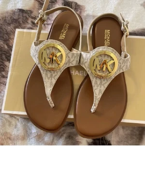 Michael Kors sandaler med gulddetalj - Snygga sandaler från Michael Kors i beige och brunt med ormmönstrad rem och stor guldfärgad MK-logga framtill. Justerbart spänne vid ankeln och platt sula i syntetmaterial. Perfekta för en trendig och avslappnad look.