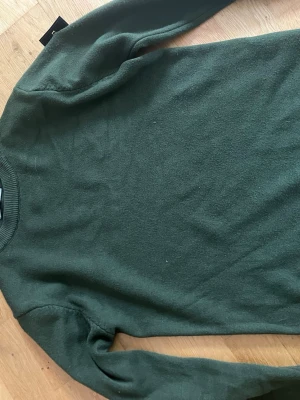 Mörkgrön stickad tröja - En stilren mörkgrön stickad tröja med rund halsringning och långa ärmar. Tröjan har en klassisk passform och ribbstickade muddar vid ärmslut och nederkant. Perfekt för lager-på-lager och enkel att matcha med jeans eller kjol.