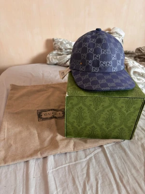 Blå GG-monogram keps från Gucci - Snygg blå keps från Gucci med klassiskt GG-monogram i vitt över hela kepsen. Justerbar rem bak i blått läder och guldfärgat spänne. Kepsen är i en klassisk baseballmodell och kommer med originalförpackning och dustbag.