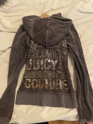  Vintage Juicy Couture grå hoodie  - Mjuk och lyxig vintage grå hoodie från Juicy Couture i velour med stor huva. Baksidan är dekorerad med glittrande guldpaljetter och text som ger en glamorös vibe. Perfekt för dig som gillar statement-plagg och vill sticka ut med en ikonisk look. 