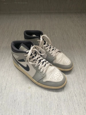 Nike Air Jordan 1 Mid grå/vit - Snygga Nike Air Jordan 1 Mid sneakers i grått och vitt med svart swoosh. Klassisk design med perforerad tåbox, vita skosnören och ikoniska Air Jordan-loggan på sidan. Ovandelen är i läder och sulan är beige. Perfekta för dig som gillar streetwear och retrostil.