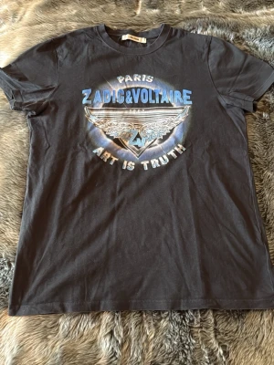 Svart ZADIG & VOLTAIRE t-shirt - Cool svart t-shirt från ZADIG & VOLTAIRE med tryck fram och bak. Framsidan har blå och silver text samt en vinge med logga, och baksidan har tour-tryck med städer och årtal. Klassisk passform och rund hals. Perfekt för dig som gillar statement-plagg.