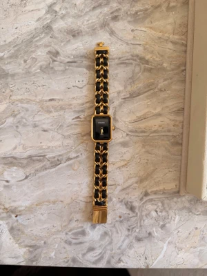Chanel svart och guld klocka - Snygg klocka från Chanel med svart urtavla och guldfärgad ram. Armbandet är en mix av svart läder och guldfärgad kedja som ger en lyxig känsla. Klassisk rektangulär form och minimalistisk design med endast visare på urtavlan. Den är liten i storlek.