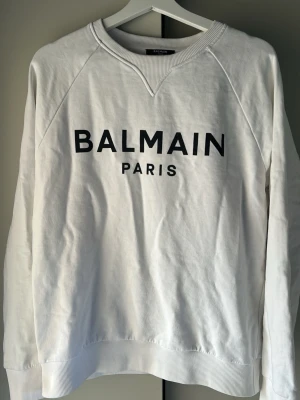 Vit sweatshirt från Balmain Paris - Snygg vit sweatshirt från Balmain Paris med svart logotyptryck på bröstet. Tröjan har rund halsringning, långa ärmar och ribbade muddar vid ärmslut och nederkant. Tillverkad i mjukt bomullsmaterial som känns skönt mot huden. Perfekt för en clean och trendig look.