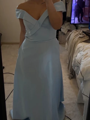 Ljusblå axelbandslös långklänning - Elegant ljusblå långklänning med offshoulder-design och vacker omlottdetalj vid bysten. Klänningen har en figurnära passform upptill och faller mjukt ner mot golvet. Perfekt för bal eller speciella tillfällen. Storlek 34