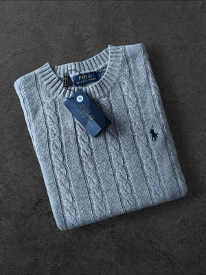 Ljusgrå kabelstickad tröja Polo Ralph Lauren - Säljer en ljusgrå kabelstickad tröja från Polo Ralph Lauren med rund halsringning och klassisk marinblå logga broderad på bröstet. Tröjan har ribbade muddar vid ärmslut och nederkant och är tillverkad i mjukt material för en skön känsla.