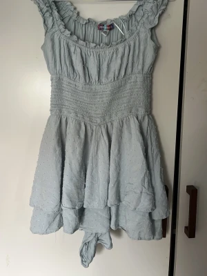 Ljusblå kort jumpsuit klänning Urban Outfitters - Supersöt ljusblå kort jumpsuit/ klänning från Urban Outfitters med volangdetaljer och smockad midja. Klänningen har ärmlös design, rundad halsringning och diskret prickigt mönster i tyget. Perfekt för varma dagar och somriga vibes.