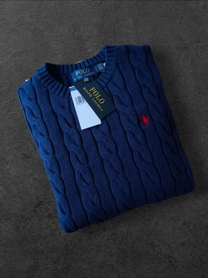 Blå kabelstickad tröja Polo Ralph Lauren - Mörkblå kabelstickad tröja från Polo Ralph Lauren med rund halsringning och röd broderad logga på bröstet. Tröjan har långa ärmar och ribbade muddar vid ärmslut och nederkant. Perfekt för dig som gillar klassisk och stilren look.