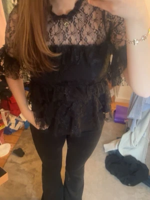 Svart spetsblus med volanger - Säljer en svart blus i spets med volangdetaljer och halvtransparent design. Blusen har korta ärmar och en feminin, romantisk look med flera lager spets. Perfekt för dig som vill sticka ut med en unik topp.