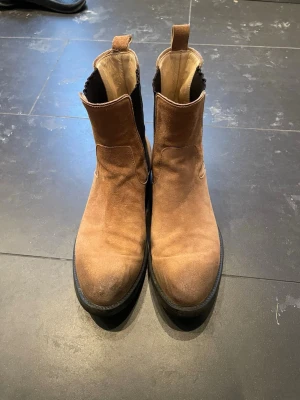 Bruna mocka chelsea boots stl 40 - Säljer ett par snygga bruna chelsea boots i mocka med svarta elastiska paneler på sidorna och dragflikar fram och bak. Skorna har platt sula och låg klack, perfekt till jeans eller chinos. Klassisk och stilren modell.