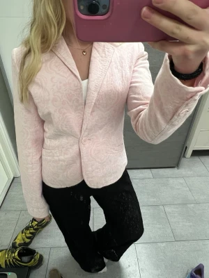 Ljusrosa mönstrad kavaj - Snygg ljusrosa kavaj med diskret mönster och klassisk krage. Kavajen har en knapp framtill och två fickor. Perfekt för att lyfta din outfit med en somrig touch. Materialet är mjukt och bekvämt och passformen är figurnära.