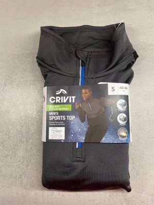 Svart sporttröja från Crivit med dragkedja - Svart sporttröja från Crivit i storlek S, tillverkad av återvunna material. Tröjan har en halv dragkedja framtill i blått och hög krage. Perfekt för träning och löpning, med funktionellt material som andas.