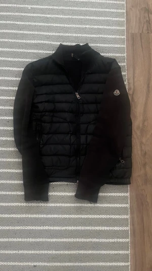ÄKTA Svart cardiganjacka från Moncler - Snygg svart cardiganjacka från Moncler med quiltad front och stickade ärmar. Jackan har dragkedja framtill, Moncler-logga på ärmen och en modern, sportig look. Perfekt för lager-på-lager och streetstyle.ÄKTA skick 8/10 solblek 