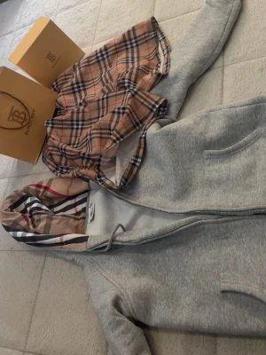 Grå Burberry hoodie med klassiskt mönster - Snygg grå hoodie från Burberry med dragkedja och huva fodrad i det ikoniska rutmönstret. Mjukt material och två fickor framtill. Perfekt för dig som vill ha en stilren och lyxig look med en touch av klassiskt brittiskt mode. Med en snygg vinter jacka moose knuckles med svart päls passar 165 till 175 längd shortsen är Xs och hoodien passar max till 164