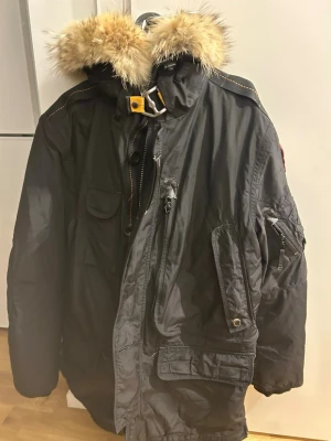 Svart parkasjacka med pälskrage - Svart parkasjacka från Parajumpers med avtagbar huva och fluffig pälskant. Jackan har flera fickor, dragkedja och knappar framtill samt gula detaljer vid hals och rygg. Perfekt för kalla dagar med sin robusta design och snygga look. Om ni har en fråga så kan ni skriva, priset är inte hugget i sten