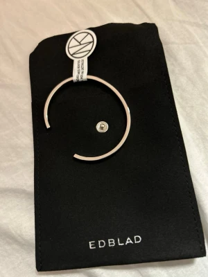 Silverfärgat armband från Edblad - Stilrent och minimalistiskt armband i silverfärg från Edblad. Armbandet är stelt och har en öppen design som gör det enkelt att ta på och av. Kommer med en svart tygpåse med Edblad-logga. Perfekt för dig som gillar enkel och modern stil. Den är använd och godi bagen är borta 🎁