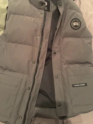 Grå frestyle crew väst Black Label Canada Goose - Snygg grå dunväst från Canada Goose med klassiska black label logga på bröstet. Köpt för flera flera år sedan. Säljer på grund av att man växt på sig några pannor och västen inte passar längre och därför att jag inte vill att den ska vara här och samla damm.