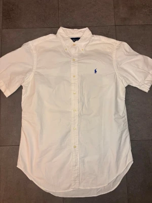 Vit kortärmad skjorta från Polo Ralph Lauren - Klassisk vit skjorta från Polo Ralph Lauren med korta ärmar och blå broderad logga på bröstet. Skjortan har knappar framtill och button-down krage. Perfekt till en clean och stilren look.