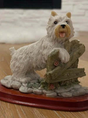 Dekorativ hundfigur  - Säljer en detaljerad porslinsfigur föreställande en vit terrierhund som lutar sig mot ett trästaket. Figurstativet är i träimitation och dekorerat med små blommor. Perfekt som prydnad till hyllan eller skrivbordet.