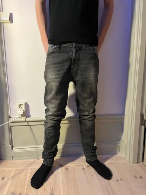 nudie  - Säljer ett par nudie  jeans i mörkgrå tvätt. Jeansen har en slim passform med raka ben och klassiska fem fickor. Tillverkade i mjukt bomullsmaterial med snygga slitningar för en modern look. använt en gång