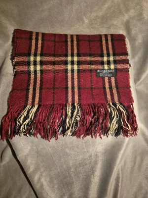 Rutig halsduk från Burberry i ull - Klassisk röd och svart rutig halsduk från Burberry i sopermjuk 100% lammull. Halsduken har fransar i ändarna och det ikoniska Burberry-mönstret med inslag av beige och gult. Perfekt accessoar för att lyfta din outfit.