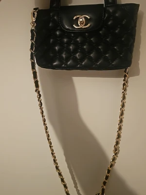 Svart axelväska i läder - Snygg svart axelväska i quiltat läder med klassiskt guldigt CC framtill. Väskan har en lång kedjerem med insytt läder och rundade handtag. Perfekt för dig som vill ha en ikonisk och trendig accessoar.