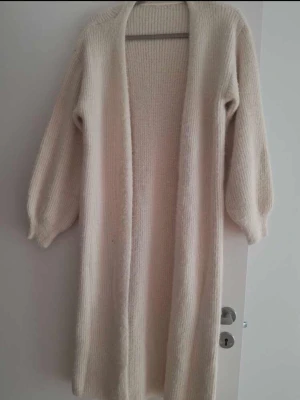 Lång beige stickad kofta - Mysig lång kofta i färgen beige. Koftan har stickat material med ribbade muddar och öppen front. Den har ballongärmar och en avslappnad passform, perfekt att slänga över en outfit för extra värme och stil.