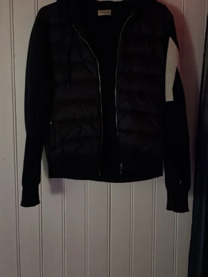 Svart Moncler jacka med stickade ärmar - Snygg svart jacka från Moncler med vadderad front och stickade ärmar. Jackan har dragkedja framtill, huva och en vit detalj på ena ärmen med Moncler-logga. Perfekt för dig som vill ha en trendig och bekväm jacka i vardagen. (Ska tvättas så alla vet)