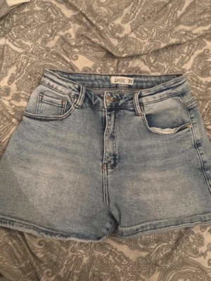 Ljusblå denimshorts från Laulia - Snygga ljusblå denimshorts från Lauli med hög midja och klassisk femficksdesign. Shortsen har bälteshällor, knapp- och dragkedjestängning samt en avslappnad passform. Perfekta för varma dagar och enkel att matcha med allt. (Köps utomlands)