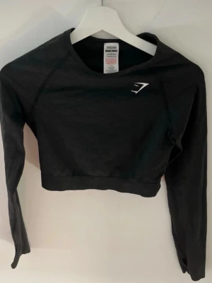 Svart crop top från Gymshark - Snygg svart långärmad crop top från Gymshark med diskret logga på bröstet. Tillverkad i ett stretchigt funktionsmaterial som andas, perfekt för träning eller gym. Tight passform och raglanärmar för maximal rörelsefrihet.