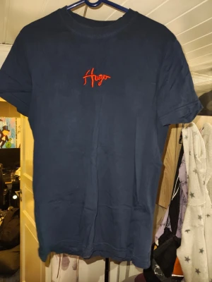 Mörkblå Hugo Boss t-shirt - Snygg mörkblå t-shirt från Hugo Boss med röd broderad Hugo-logga på bröstet. Klassisk rund halsringning och korta ärmar. Tillverkad i mjuk bomull som känns skön mot huden. Perfekt för en clean och stilren look.