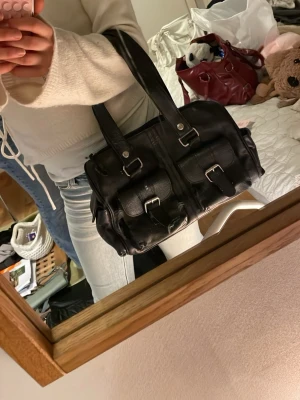 Svart handväska med spännen - Snygg svart handväska i skinnimitation med två framfickor och silverfärgade spännen. Väskan har dubbla handtag och en cool, lite boxig form som rymmer det mesta du behöver. Perfekt för dig som gillar en edgy och praktisk look.