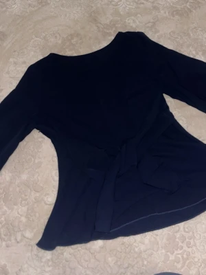 Mörkblå topp med knyt i midjan - Snygg mörkblå topp med långa ärmar och trendigt knyt i midjan. Perfekt för dig som gillar stilrena plagg med en twist. Materialet känns mjukt och följsamt, vilket gör toppen bekväm att bära. Enkel att matcha med både jeans och kjol.