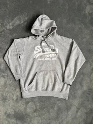 Grå SUPERDRY Hoodie - Säljer en fin grå hoodie från Superdry. Hoodien är i använt skick. Snörena är lite slitna längst ner och loggan är lite sliten annars är allt annat bra. Storlek L. Hör av dig vid minsta fundering!😊