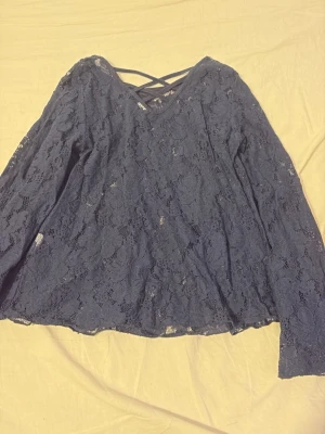 Mörkblå spetsblus med korsad rygg - Snygg mörkblå blus i spets med långa ärmar och v-ringning. Baksidan har en trendig korsad detalj vid nacken. Blusen är genombruten och har en feminin, avslappnad passform. Perfekt att styla med jeans eller kjol för en cool look.